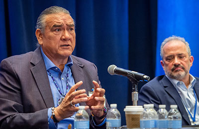 A man with long grey hair pulled back speaking into a tabletop microphone with another man looking on.